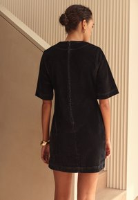 Crna traper haljina s kratkim rukavima, ravnog kroja i zatvaračem straga. Ima kontrastne šavove i glatku teksturu.