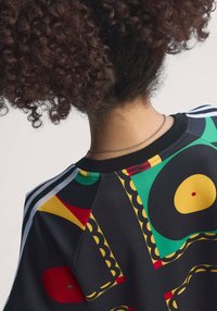 Person mit lockigem Haar, die ein bunt gemustertes Hemd mit schwarzen, gelben, roten, grünen Formen und weißen Streifen auf der Schulter trägt.