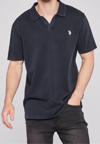 Granatowa koszulka polo wykonana z miękkiego materiału, z dekoltem w kształcie litery V i krótkimi rękawami. Małe haftowane logo znajduje się na lewej piersi.