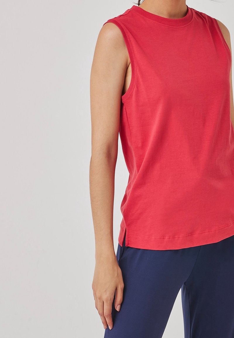 Haut rouge sans manches avec encolure ronde porté par une personne au bras détendu le long du corps, associé à un pantalon bleu marine sur un fond clair uni.