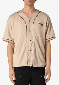 Camicia beige a maniche corte con bottoni e rifiniture nere, realizzata in tessuto perforato, con un logo cucito sul lato sinistro del petto.