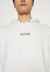 Baltas megztinis su gausia medžiaga, pasižymintis kengūros kišene, raišteliais su metaliniais akcentais ir juodu "GUESS" logotipu ant krūtinės.