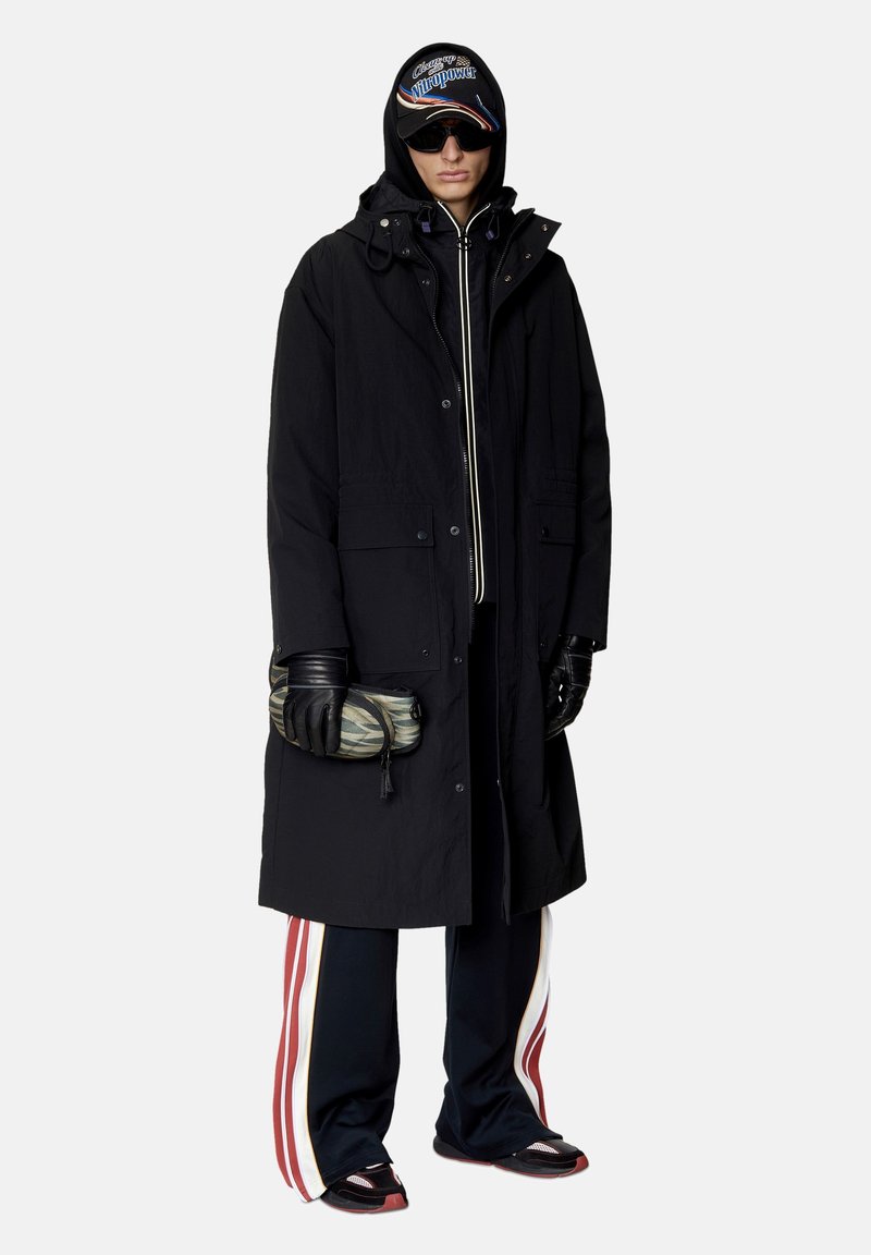 Persoon in een zwarte gewatteerde coat met capuchon, zonnebril, zwarte handschoenen, gestreepte joggingbroek, sneakers en met een tas met zebra-strepen in de hand.
