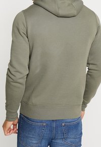 Hoodie vert olive en tissu texturé, avec capuche à cordon de serrage et poignets côtelés. Porté avec un jean bleu, montrant un dos décontracté.