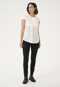 White blouse with short sleeves and pleated detailing, paired with slim black pants and black shoes featuring a chunky sole. Plain background.