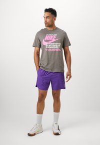 Graues Nike-T-Shirt mit pinkem Logo und Text, kombiniert mit lila Shorts. Das Model trägt weiße Socken und mehrfarbige Sportschuhe.