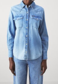 Světle modrá džínová košile s dlouhým rukávem a patentovými knoflíky, s špičatým límečkem a dvěma kapsami na hrudi. Obsahuje kontrastní stehy a zakřivený lem.