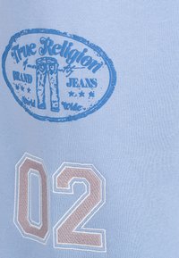 Hellblauer Stoff zeigt ein rundes Logo in dunklerem Blau mit dem Schriftzug "True Religion Brand Jeans" und einer numerischen "02" in beigem Zierstich.