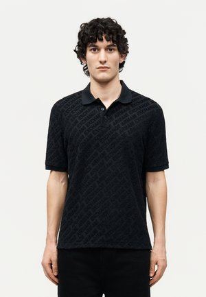 Άνδρας με σγουρά μαλλιά φοράει μαύρο κοντομάνικο πόλο με διακριτικό διαγώνιο μοτίβο κειμένου "Armani Exchange" σε ανοιχτό φόντο.