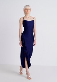 Marineblauwe mouwloze jurk met een getailleerde snit, een ruffle-detail aan de zijkant en een high-slit tot boven de dij, gemaakt van een gladde stof. Zilveren hakken.