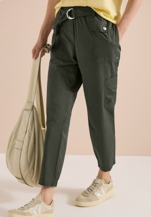 Osoba nosząca czarne spodnie typu cargo z paskiem, beżowe trampki, trzymająca beżową torbę, stojąca na tle jednolitego jasnego tła.