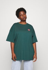 Donkergroene katoenen oversized t-shirt met een ronde halslijn en korte mouwen, voorzien van een roze bloemenontwerp op het linkerborstgebied.