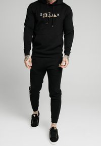 Felpa nera con logo dorato, abbinata a pantaloni jogger neri coordinati e sneakers nere con occhielli dorati. Tessuto morbido, design aderente.