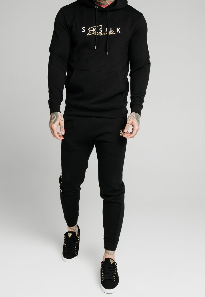 Felpa nera con logo dorato, abbinata a pantaloni jogger neri coordinati e sneakers nere con occhielli dorati. Tessuto morbido, design aderente.