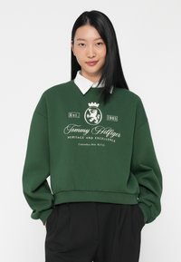 Felpa verde con maniche lunghe, colletto rotondo e orlo a coste. Presenta una stampa grafica bianca con uno stemma di leone e dettagli testuali.