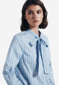 Blusa azzurro chiaro con sottili strisce bianche, caratterizzata da un collo con nodo, maniche lunghe e un tessuto liscio e lucido. Bottoni visibili sulla parte anteriore.