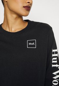 Fekete, hosszú ujjú pamut póló, amelyen egy kicsi fehér "Huf." logó található a mellkason, valamint a "Huf World" felirat fehér színben az ujján.
