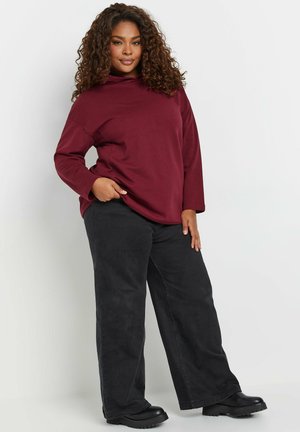 Haut à capuche bordeaux avec col montant, coupe décontractée et manches longues, associé à un pantalon large noir et des bottes noires épaisses.