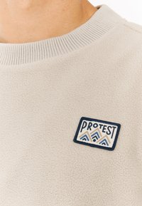 Lichtbeige fleece sweatshirt met ribgebreide kraag. Voorzien van een blauw en beige geborduurd logo patch met bergontwerp aan de voorkant.