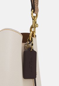 Valkoinen nahkabagi, jossa on oranssia ommeltu, varustettu ruskealla nahkaleimalla, kultaisilla hardware-osilla ja teksturoidulla pinnalla.