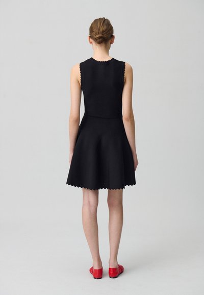 Robe sans manches noire avec une jupe évasée et des bords festonnés. Associée à des chaussures rouges, vue de dos sur un fond neutre.