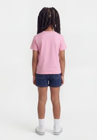 Παιδί με μακριά πλεγμένα μαλλιά που φοράει ροζ μπλουζάκι με κοντά μανίκια, σκούρο τζιν σορτς, λευκές κάλτσες και λευκά αθλητικά παπούτσια, στέκεται με την πλάτη προς τα εμπρός.