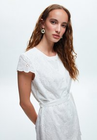 Bílé vyšívané šaty s obláčkovým výstřihem, krátkými rukávy a dekorativními knoflíky. Mají zavázaný pas a květinový perforovaný vzor.
