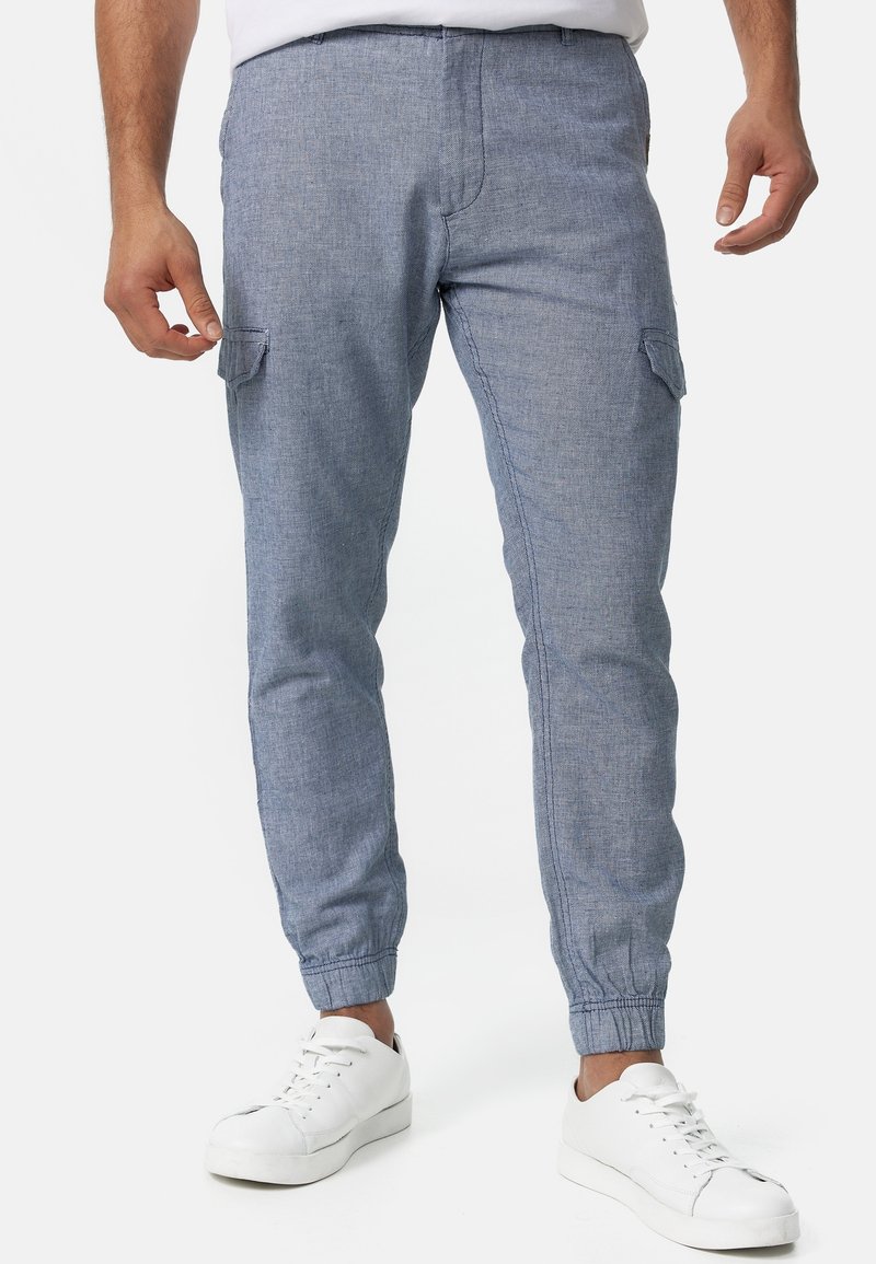Pantalons cargo bleu clair en tissu texturé avec des poignets élastiques et une poche latérale. Portés avec des baskets blanches.