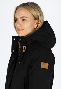 Black waterproof jacket with a hood, featuring a tan leather accent patch, zip closure, and drawstring around the hood for adjustment.