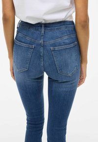 Jeans skinny en denim bleu foncé, avec une taille haute, des poches arrière et une texture lisse. Les coutures sont visibles sur les bords.