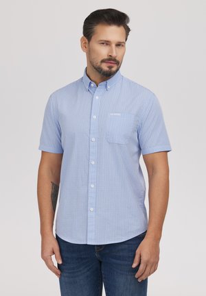 Mężczyzna z ciemnymi włosami i brodą, ubrany w jasnoniebieską krótki rękaw koszulę w kratę zapinaną na guziki oraz niebieskie dżinsy, stojący na jednolitym tle.