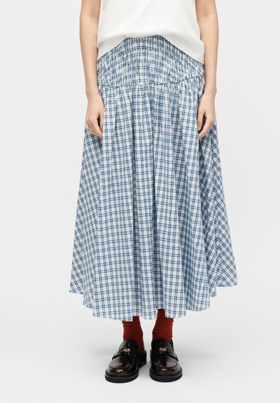 Γυναίκα που φοράει φούστα με μπλε και λευκό καρό με ψηλή μέση, κόκκινες κάλτσες, μαύρα λουστρίν παπούτσια και λευκό τοπ, όρθια μπροστά σε απλό φόντο.