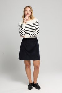 Camisa de manga larga color crema con rayas marinas, combinada con una falda A-line de ante azul marino. La modelo lleva mocasines negros, con los brazos cruzados, en un fondo neutro.