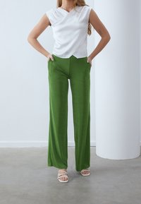 Witte mouwloze top met een V-hals ontwerp, gecombineerd met groen high-waisted wijde broeken. Model draagt witte strap-sandalen. Eenvoudige achtergrond.