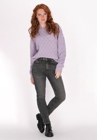 Lila kötött pulóver gyémánt mintával, magas derékú sötétszürke farmernadrággal és fekete vastag bokacsizmával párosítva. Semleges háttér.