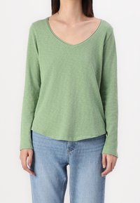 Maglia a maniche lunghe di colore verde chiaro, realizzata in tessuto morbido. Presenta un profondo scollo a V e una vestibilità rilassata con orlo curvo. Abbinata a jeans azzurri.