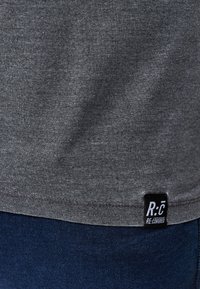 Šedé bavlněné tričko se hladkým povrchem a černou cedulkou na spodním lemu se nápisem "R:C RE-COVERED". Námořnické džíny viditelné níže.