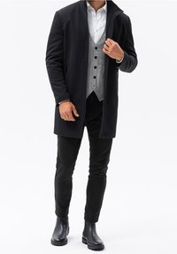 Svart lång kappa, grå väst med knappar, vit skjorta, svarta byxor och blanka svarta ankelboots. Texturerad tyg med rena linjer och en åtsittande form.