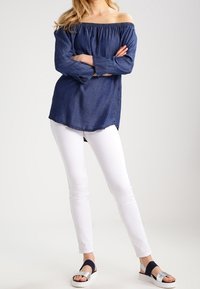 Femme portant un haut bleu à épaules dénudées, un pantalon blanc ajusté et des sandales argentées à bouts ouverts avec des brides noires, debout les bras croisés.