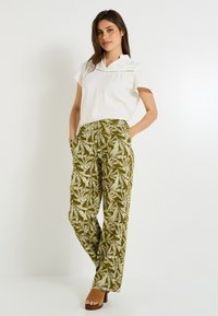 Chemise blanche à manches courtes avec des détails en dentelle, associée à un pantalon ample vert à motifs, arborant des motifs de feuilles. Chaussures à plateforme marron.