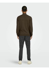 Suéter de punto marrón con cuello alto, combinado con pantalones de cuadros grises. El modelo lleva zapatillas de color claro, visto desde atrás.