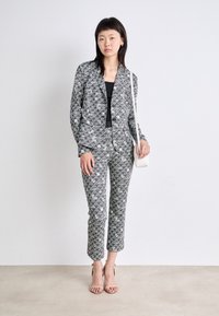 Schwarz-weiß gemusterter taillierter Anzug mit einem Kragenhemd und schmal zulaufenden Hosen. Modell hält eine kleine weiße Tasche. Betont mit zarten High Heels.