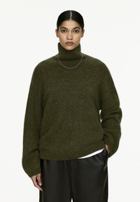 Dark green turtleneck sweater made of soft, textured material, with a relaxed fit and dropped shoulders, paired with black pants.