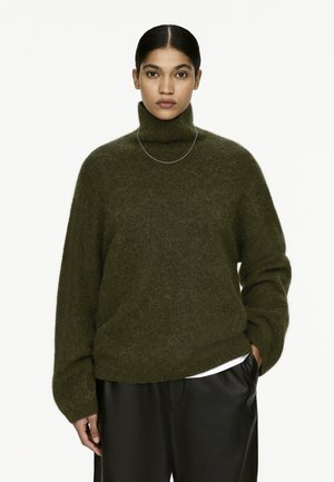 Dunkelgrüner Rollkragenpullover aus weichem, strukturiertem Material, mit lässiger Passform und überschnittenen Schultern, kombiniert mit schwarzen Hosen.