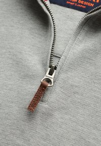 Maglione grigio con zip, realizzato in tessuto texturizzato, caratterizzato da una cerniera in metallo argentato e un'etichetta in pelle marrone con il logo "Superdry" impresso.