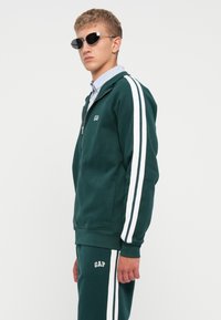 Green zip-up tracksuit with white stripes on sleeves and pants. Made of soft fabric, featuring a logo on the chest and waist.