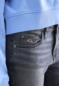 Sudadera azul acanalada combinada con jeans oscuros. Los jeans tienen un pequeño parche de logo en el bolsillo y detalles metálicos en los botones.