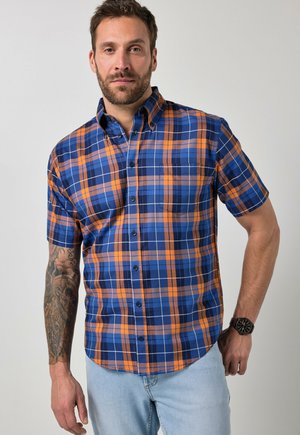 Mann mit Bart, der ein kurzärmeliges blau-orange kariertes Hemd und helle Jeans trägt, zeigt ein Tattoo am linken Arm und trägt eine schwarze Armbanduhr.