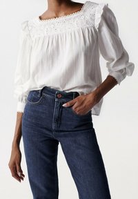 Weiße Bluse aus strukturiertem Stoff mit Rüschensäumen und gesticktem Ausschnitt, kombiniert mit dunkelblauen Jeans mit Knopf und klassischer Passform.