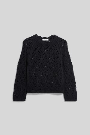 Red-haired woman wearing a black, long-sleeve, open-knit sweater and a black skirt, standing against a plain light background.